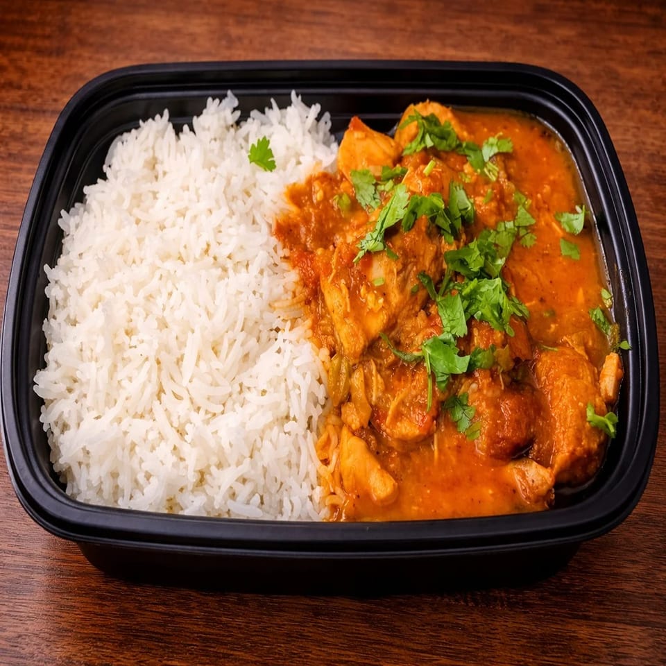 Chicken curry with rice and cilantro in a black to-go container on a wooden table.