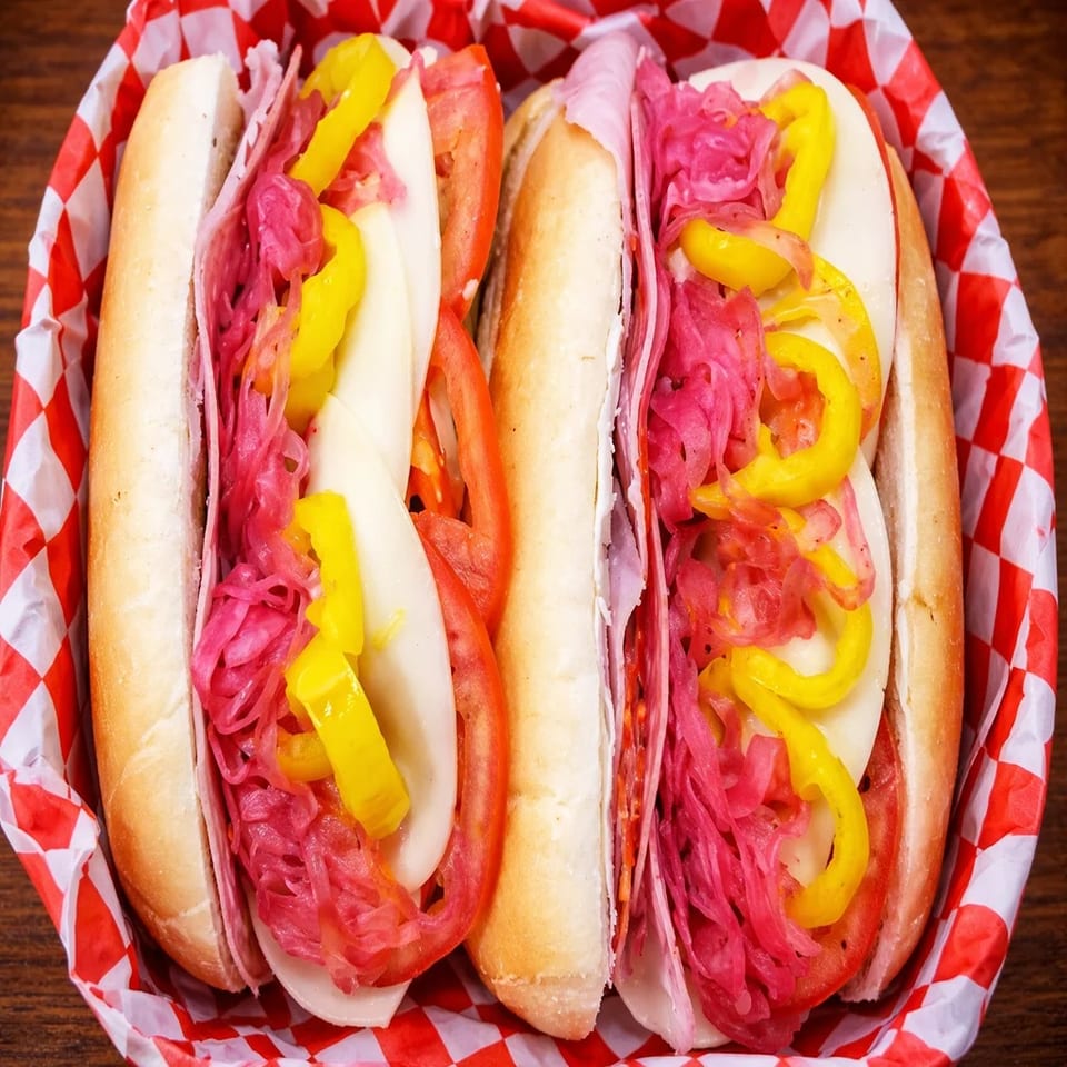 Two colorful subs with meat, cheese, tomatoes, yellow peppers, and red pickled onions in a red checkered basket.